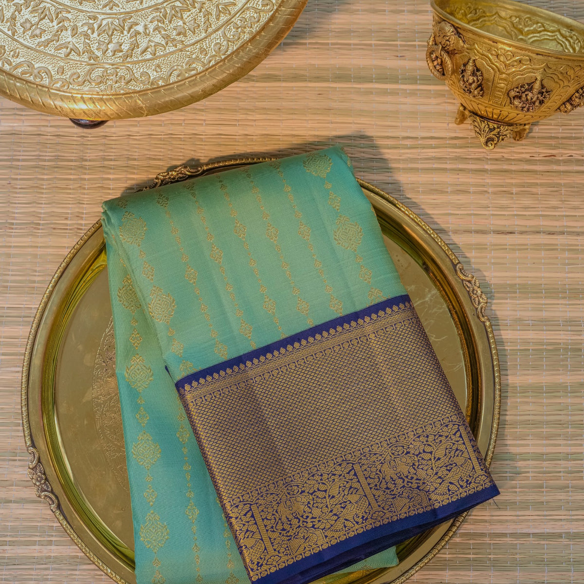 A folded turquoise blue saree with gold zari motifs and a dark blue border, displayed on an ornate gold-toned plate over a woven mat.