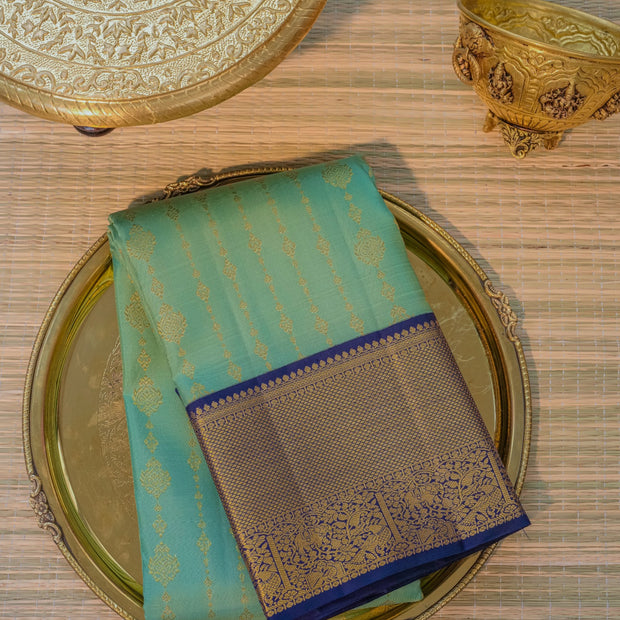 A folded turquoise blue saree with gold zari motifs and a dark blue border, displayed on an ornate gold-toned plate over a woven mat.